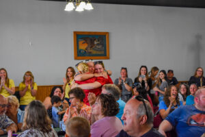 A crowded room of cheering spectators, with two women embracing in the foreground while others clap and smile.